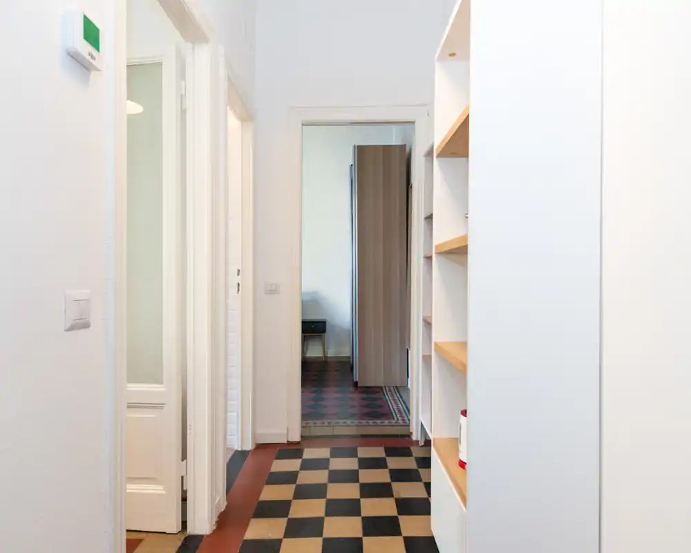 Pasillo de entrada luminoso con suelo ajedrezado clásico, estanterías empotradas a la derecha y puerta hacia las habitaciones interiores. Paso limpio y bien iluminado, perfecto para destacar la circulación en un anuncio.