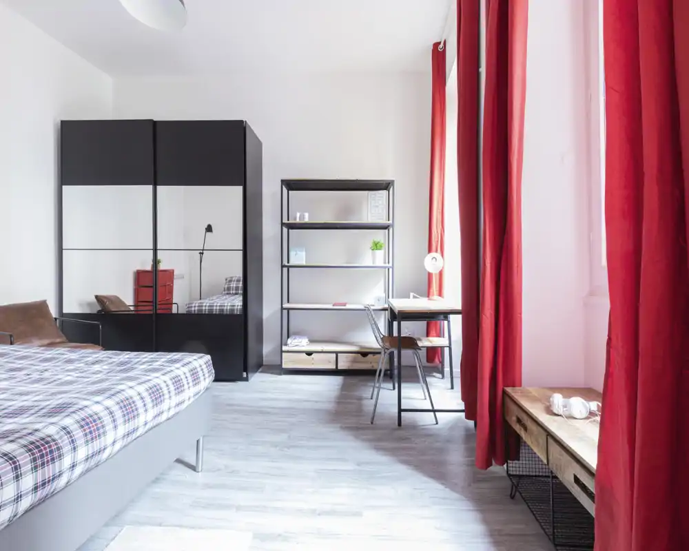 Camera da letto moderna e luminosa con letto a quadri a sinistra, armadio con specchi, mensole aperte e una scrivania vicino a una grande finestra con tende rosse vivaci.