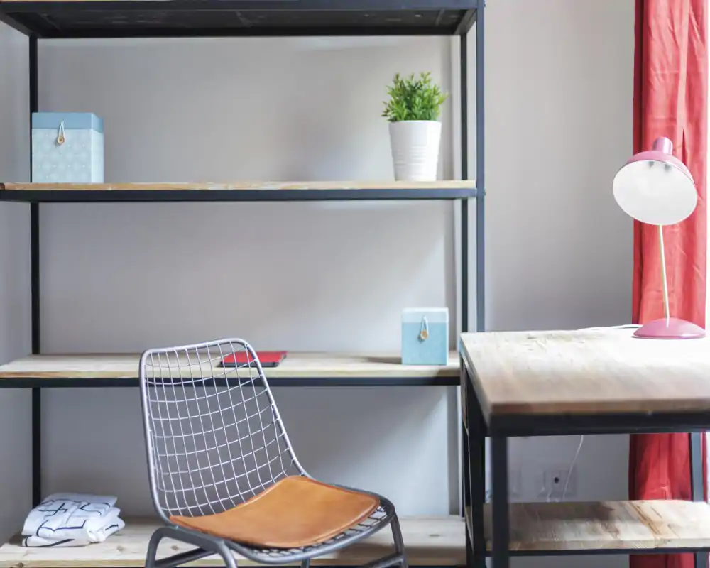 Angolo soggiorno minimalista e luminoso con scaffalatura in metallo, sedia a rete con seduta in pelle, scrivania in legno e lampada da tavolo rosa — stile semplice e funzionale, ideale per uno studio moderno.