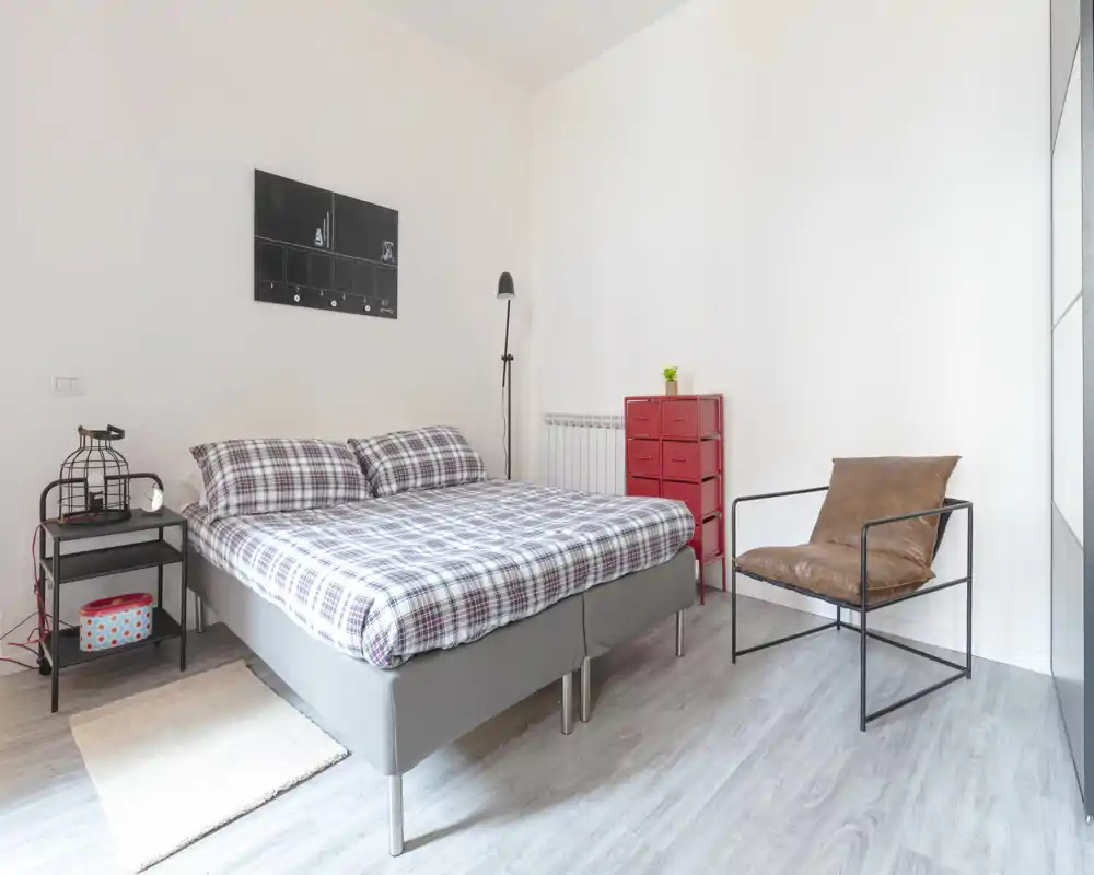 Camera da letto moderna e luminosa con un letto matrimoniale ben rifatto, biancheria a quadri, comodino con lampada e una comoda poltrona: ideale per annunci in affitto che valorizzano uno spazio pulito e minimale.