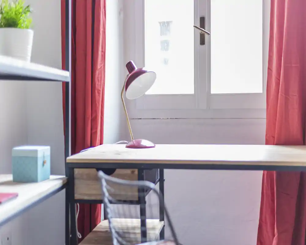 Angolo lavoro luminoso con scrivania in legno, sedia in metallo e lampada rosa sotto una grande finestra; tende rosse e una piccola pianta regalano colore.