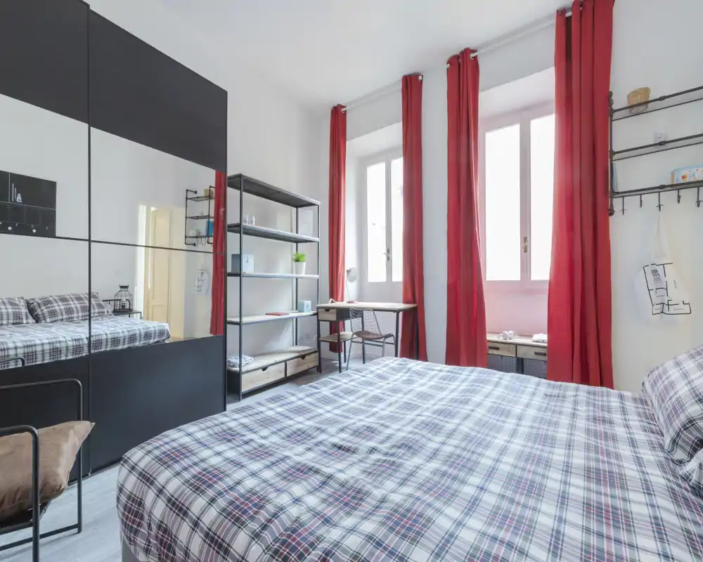 Camera da letto luminosa e ben composta con un ampio letto in primo piano, tende rosse e un guardaroba con specchio che aggiunge profondità. Presente una scrivania e scaffali aperti, ideale per un soggiorno confortevole e funzionale.