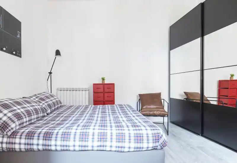 Camera da letto moderna e luminosa con un ampio letto con copriletto a quadri, una poltrona in pelle e un armadio con ante specchiate che ampliano lo spazio visivo.