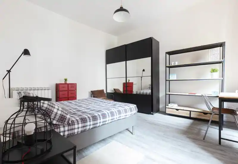 Camera da letto moderna e luminosa con letto matrimoniale con copertura a quadri, ampia armadiatura con specchi, scaffalatura aperta e una scrivania ordinata — stile pulito e tanta luce naturale.
