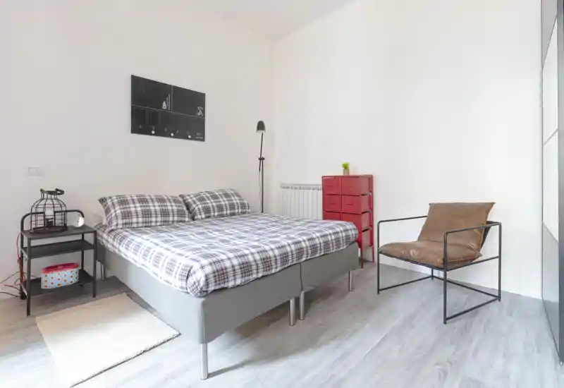 Bright, modern bedroom with a neatly made double bed, patterned bedding, a bedside table with lamp and a comfortable accent chair — ideal for rental photos highlighting a clean, minimalist sleeping space.