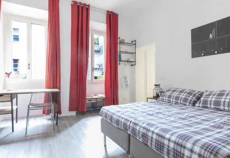 Camera da letto moderna e luminosa con letto matrimoniale con copriletto a quadri, ampie finestre con tende rosse, una piccola scrivania e mensole ordinate — perfetta per soggiorni brevi e confortevoli.