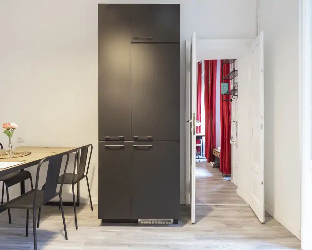 Angolo cucina/pranzo moderno con un alto armadio scuro e un piccolo tavolo con sedie in metallo; luminoso, minimale e ben arredato per un appartamento compatto.