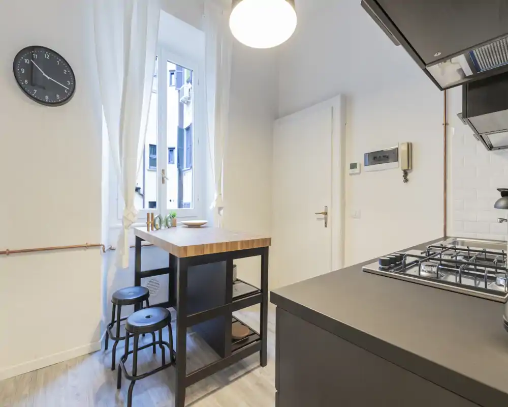 Cucina compatta e moderna con isola colazione in legno, due sgabelli e piano cottura a gas integrato. Pareti bianche e tende creano un’atmosfera luminosa e ariosa, perfetta per appartamenti urbani.