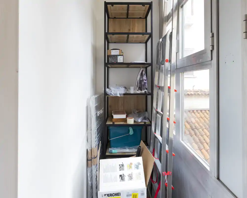 Stretto spazio tipo balcone usato come ripostiglio con scaffalature, scatole e una scala vicino a grandi finestre che lasciano entrare luce naturale.