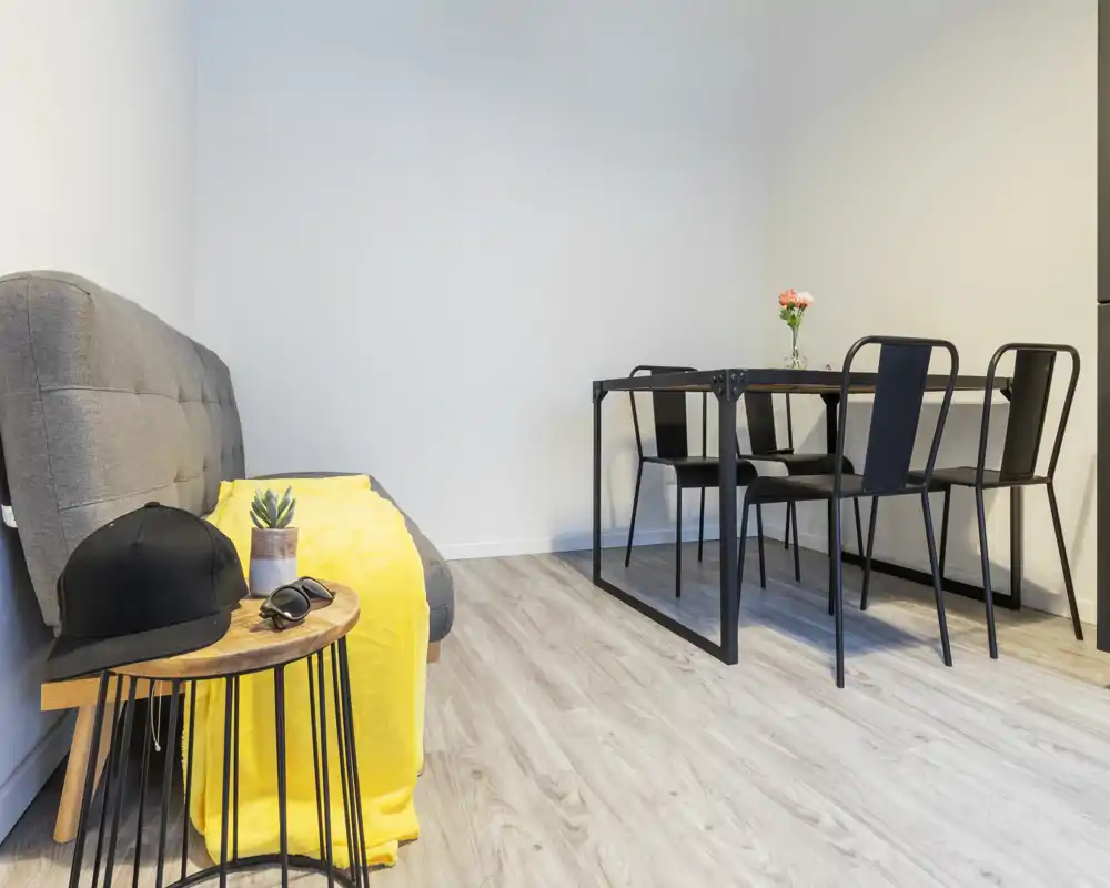 Zona giorno luminosa e minimal con divano grigio compatto, plaid giallo e un piccolo tavolo da pranzo con quattro sedie nere — ideale per un monolocale moderno.