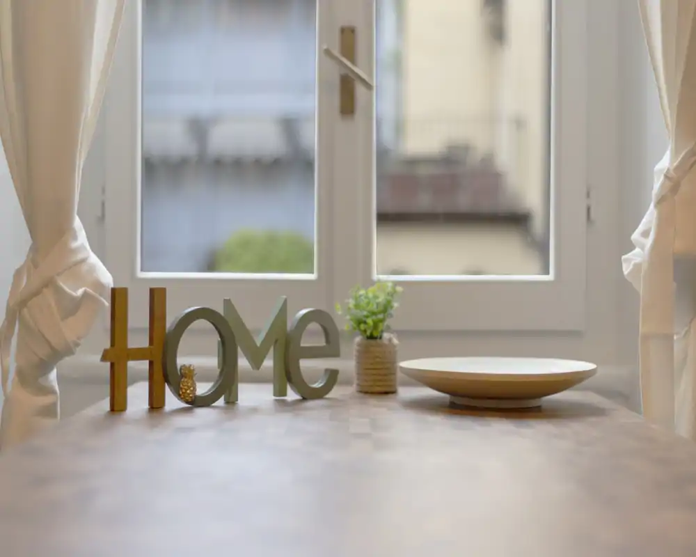 Tavolo in stile vicino a una finestra luminosa con decorazioni 'HOME', una piccola pianta in vaso e un vassoio in legno—perfetto per mostrare l'arredo da tavola e la luce naturale.