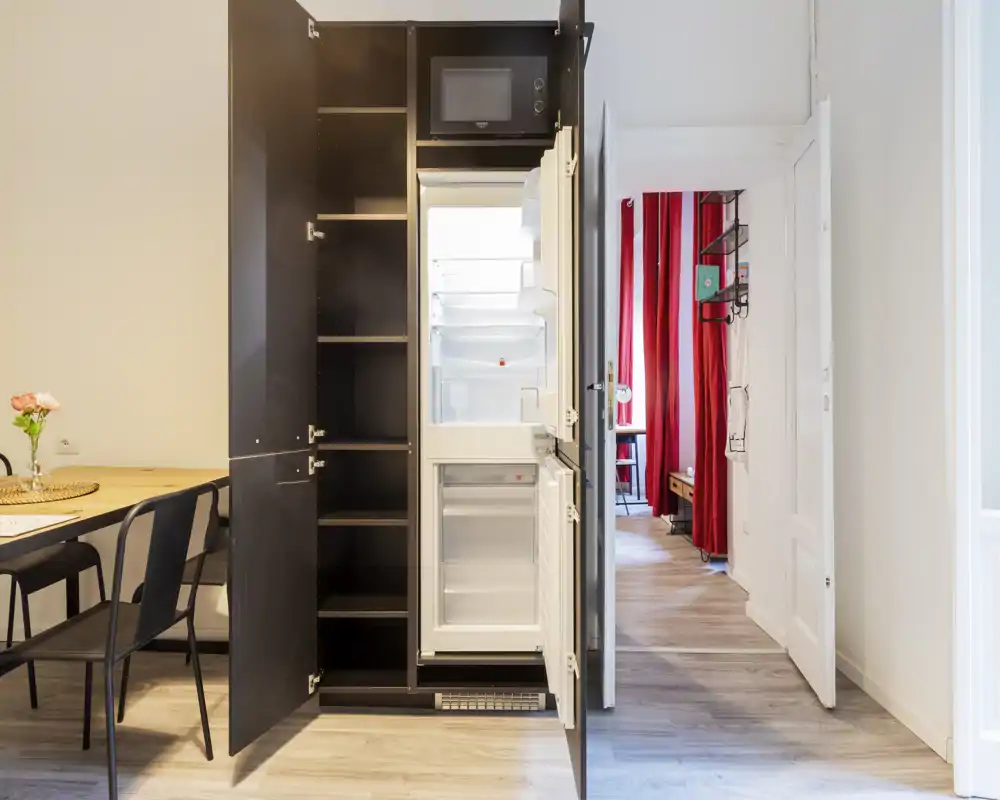 Angolo cucina compatto con frigorifero integrato in mobili scuri, piccolo tavolo con sedie e pavimento in legno chiaro; luminoso, ordinato e pronto all'uso.