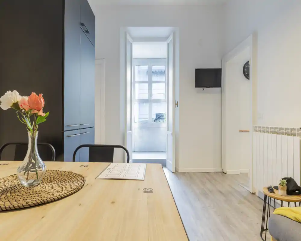 Zona giorno luminosa e moderna con un grande tavolo in legno in primo piano, vaso con fiori, sedute e una TV a muro. La luce naturale entra dalla finestra incassata, creando un ambiente accogliente e ben arredato, ideale per cenare e rilassarsi.