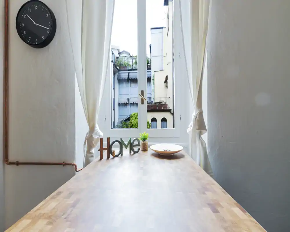 Angolo colazione luminoso con piano in legno, decorazione "HOME", una piccola pianta e un piatto, incorniciato da una grande finestra che lascia entrare luce naturale.
