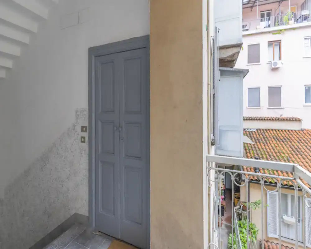 Piccolo ingresso/androne con porta doppia verniciata e vista dal balcone sui tetti in tegole. La luce naturale entra da destra, creando un ambiente di transizione accogliente.