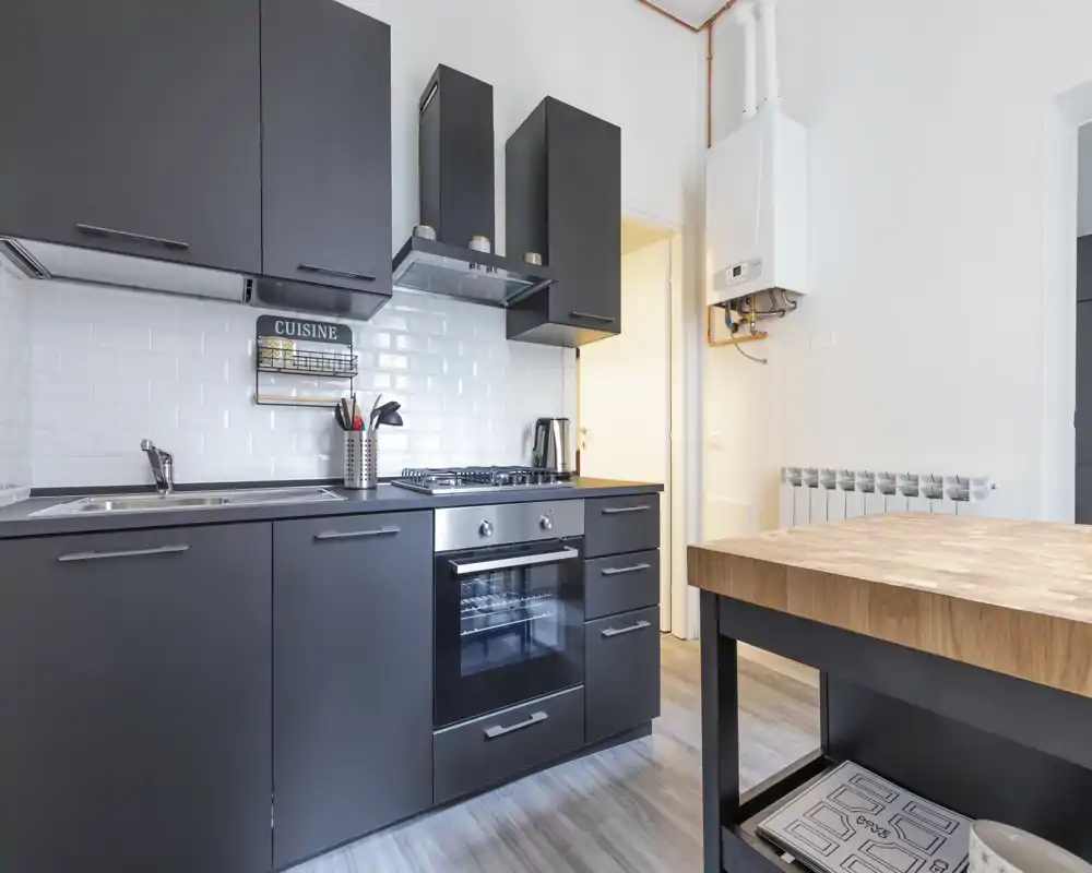 Cucina compatta e moderna con pensili scuri opachi, forno incassato, piano cottura a gas e isola con top in legno. Schienale con piastrelle subway bianche e buona luce naturale per un ambiente funzionale.