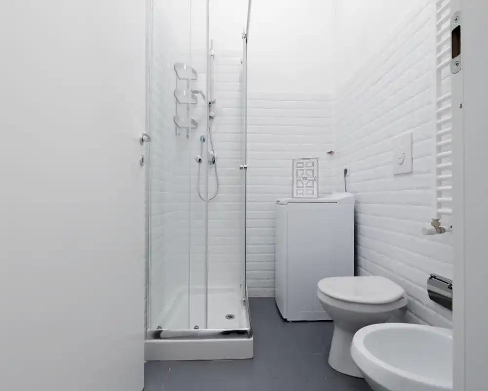 Bagno moderno e luminoso con box doccia in vetro, WC e bidet su pavimento piastrellato; mattonelle bianche e una lavatrice compatta rendono lo spazio funzionale e ordinato.