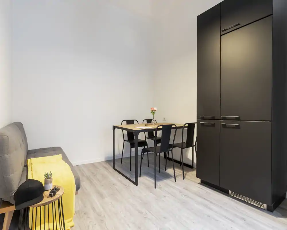 Piccolo soggiorno moderno con divano compatto, tavolo da pranzo per quattro e armadio alto nero opaco; luminoso, minimale e ben arredato per annunci.