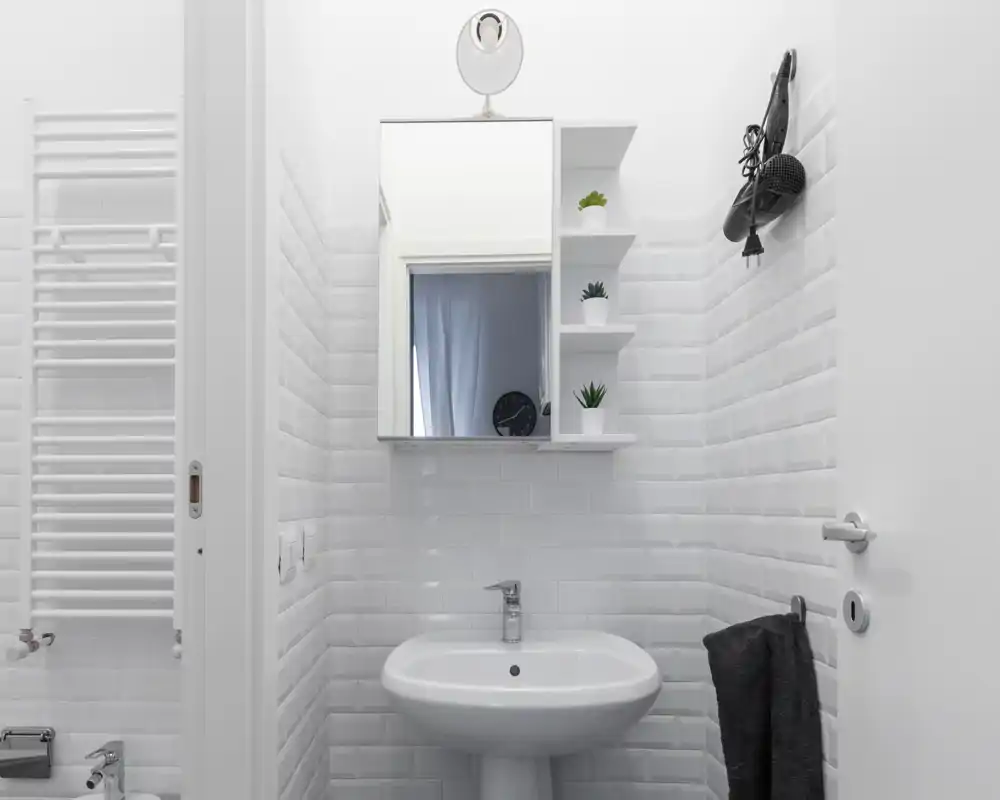 Bagno moderno e luminoso con piastrelle bianche, lavabo su colonna, specchio con armadietto e mensoline: ambiente pulito, essenziale e ben illuminato.