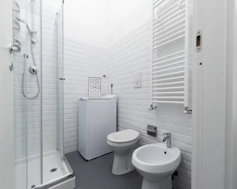 Bagno compatto e moderno con cabina doccia in vetro, wc, bidet e una piccola lavatrice. Piastrelle bianche pulite e sanitari contemporanei per un aspetto fresco e minimal.