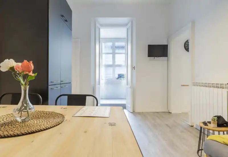 Bright, modern living area with a large wooden table in the foreground, decorative vase with flowers, seating and a TV mounted on the wall. Natural light floods the space through a recessed window, creating a welcoming, well-styled common room ideal for dining and relaxing.