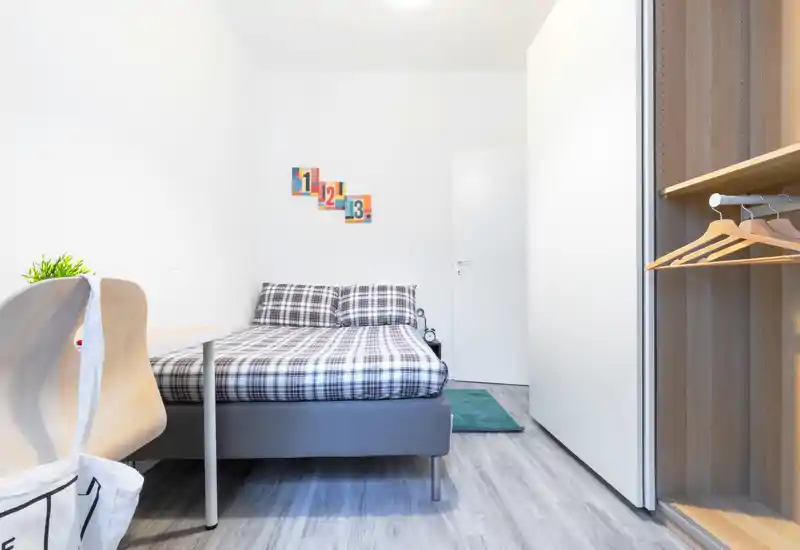 Bright, minimal bedroom with a double bed in plaid bedding, a small desk and an open wardrobe. The space feels clean and well lit, ideal for a rental listing highlighting comfort and storage.