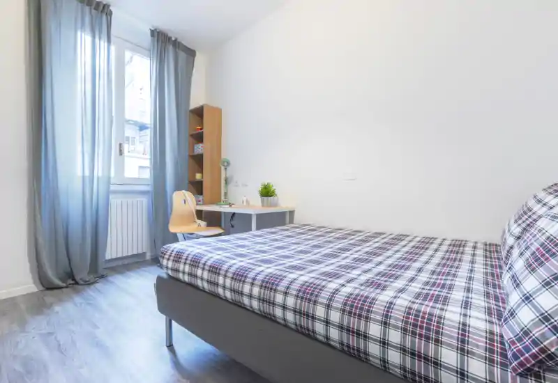 Bright, modern single bedroom with a comfortable bed dressed in plaid bedding, a small desk and shelving by the window, and soft natural light.
