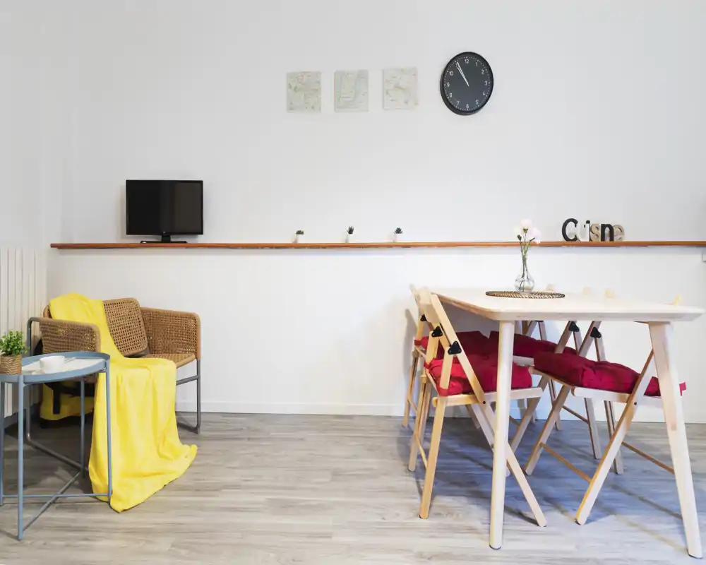 Bright, minimalist living/dining area with a small table and folding chairs, a rattan armchair with a yellow throw, and simple wall decor — suitable for cozy short-term stays.