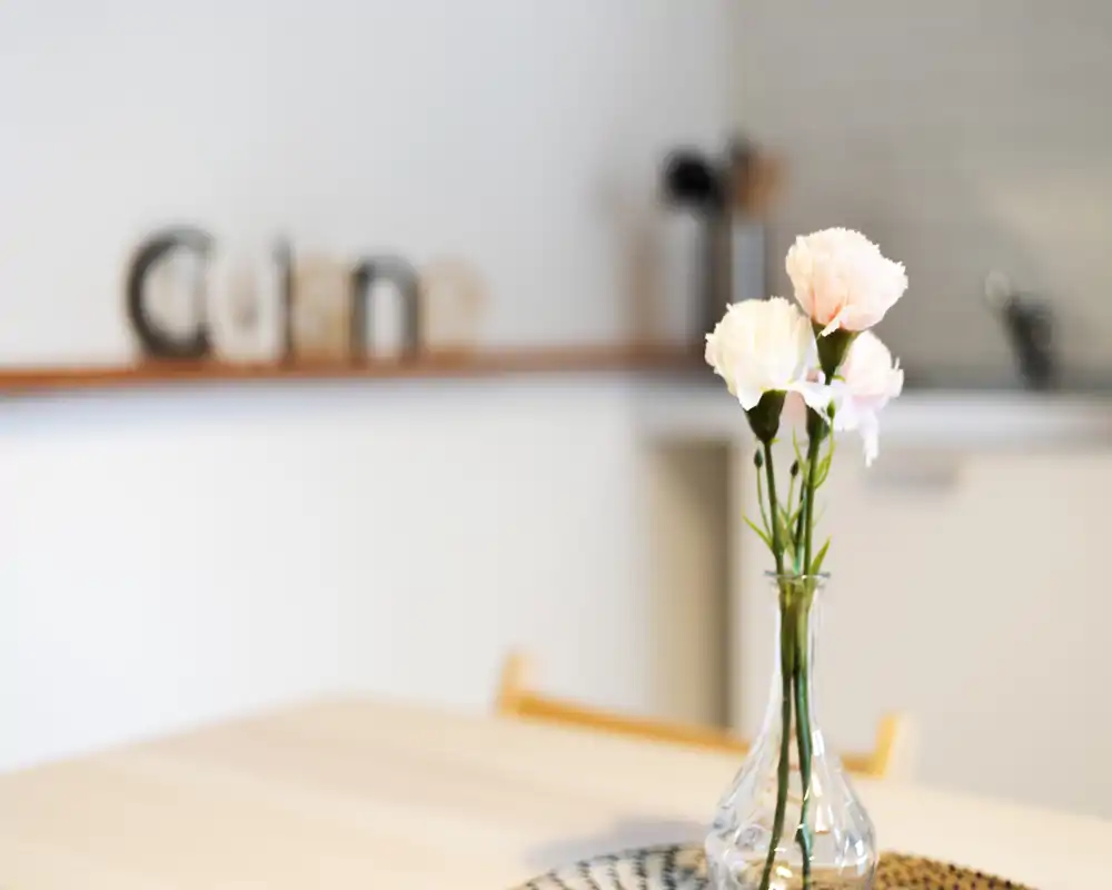 Close-up in a bright kitchen showing a decorative glass vase with fresh flowers on a wooden table; the countertop and kitchen details are softly blurred in the background, creating a warm, styled look.