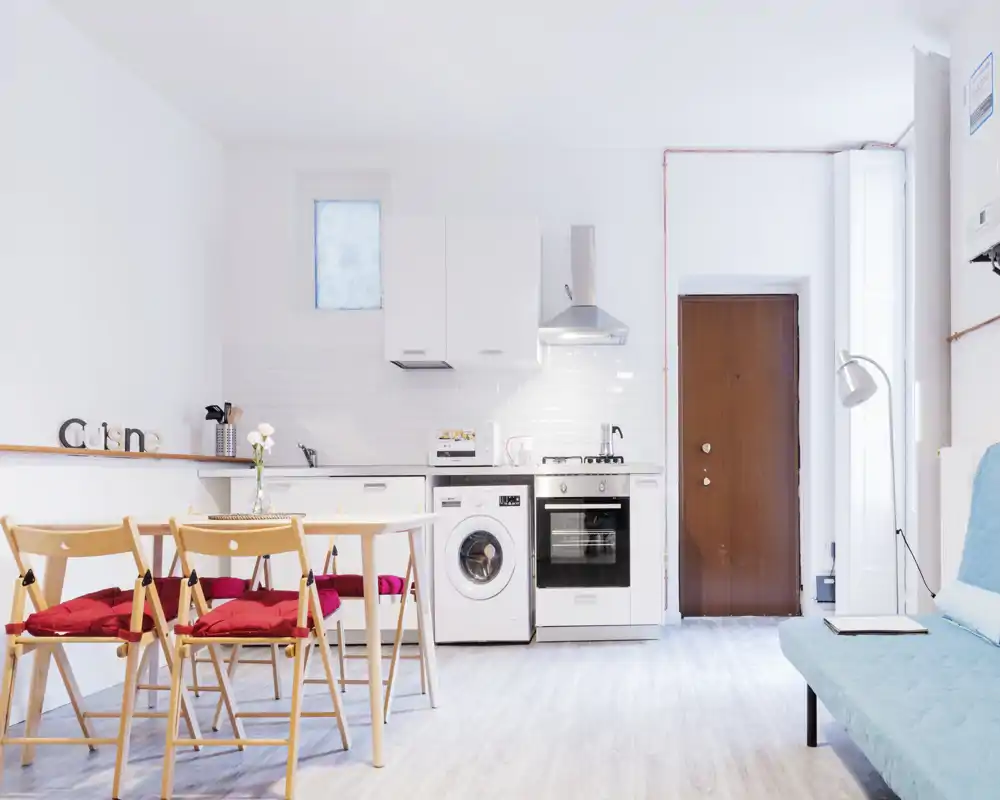 Bright open-plan kitchen with built-in appliances, a washer and oven, a small dining table with red-cushioned chairs and minimalist white cabinetry — ideal for compact apartments.