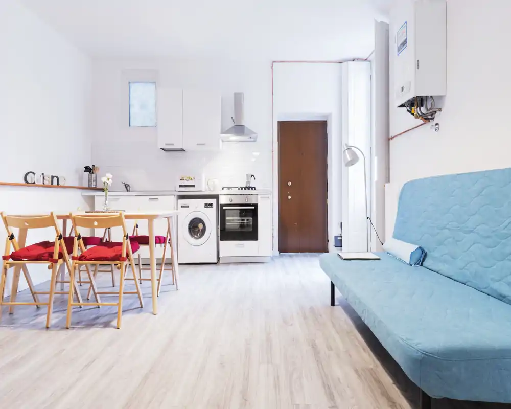 Bright, modern open-plan living area with a compact kitchen and a blue sofa. Light wood flooring and red-cushioned folding chairs give a fresh, airy feel ideal for short-term rentals.