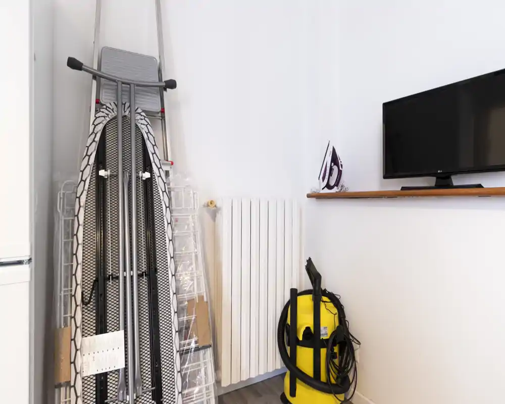 Small utility corner or entry area used for storage: ironing board, ladder, vacuum cleaner and a wall-mounted TV on a simple shelf. Clean white walls and laminate flooring with natural light.