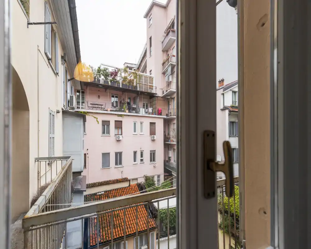 View from a small urban balcony overlooking neighboring apartment buildings and a tiled roof; narrow exterior railings and a window handle are visible in the foreground.