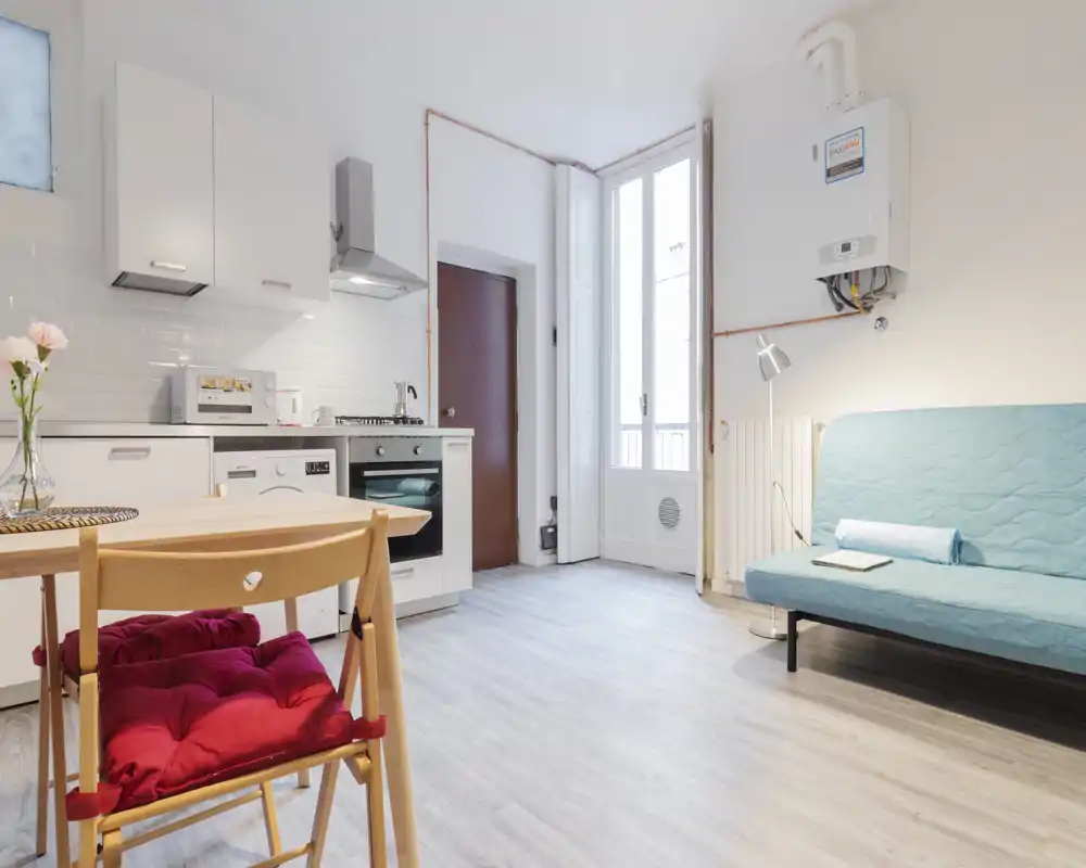 Bright, modern studio-style living area with a compact kitchenette, dining chair with cushion, and a teal sofa—ideal for a small apartment rental.