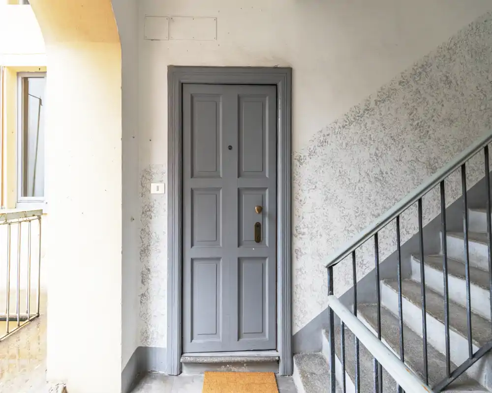Welcoming apartment entrance with a painted door, doormat and adjacent stairwell; bright natural light and textured walls give a neat, lived-in feel.