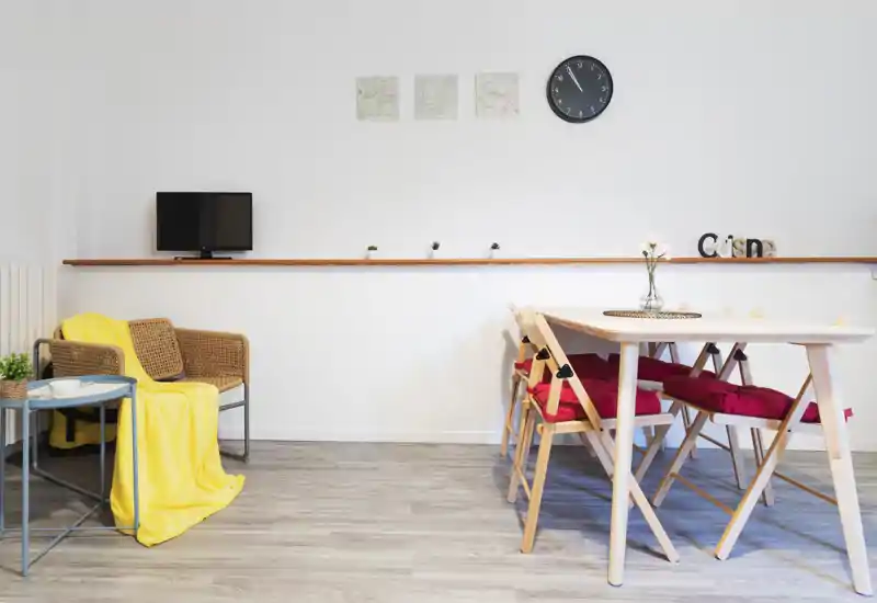 Bright, minimalist living/dining area with a small table and folding chairs, a rattan armchair with a yellow throw, and simple wall decor — suitable for cozy short-term stays.