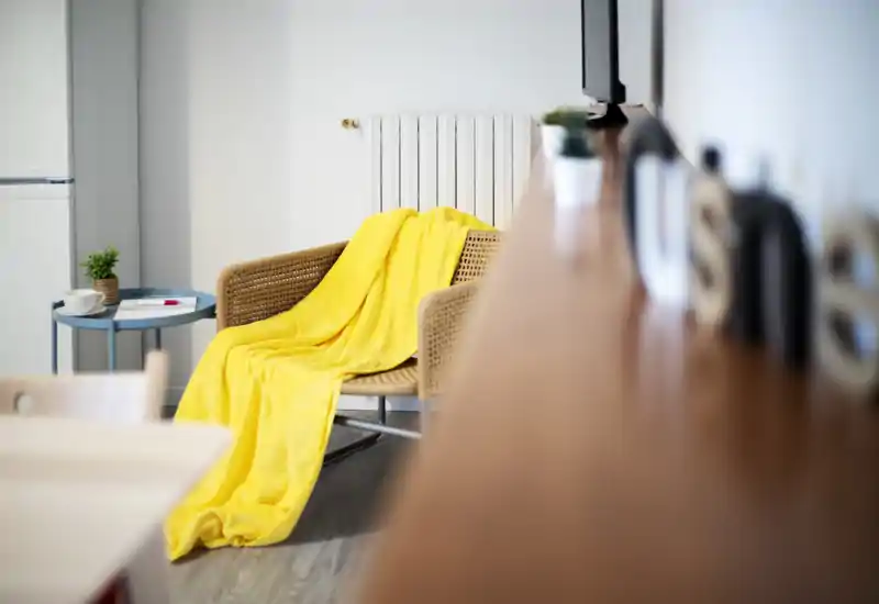 Bright, modern living corner with a woven armchair draped in a vibrant yellow throw, a small side table with a plant and cup, and soft, clean lighting.