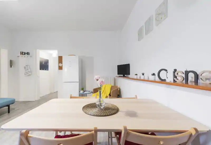 Bright, minimalist dining area with a wooden table set, fresh flowers centerpiece and cozy accents like a yellow throw and cushions creating an inviting atmosphere.
