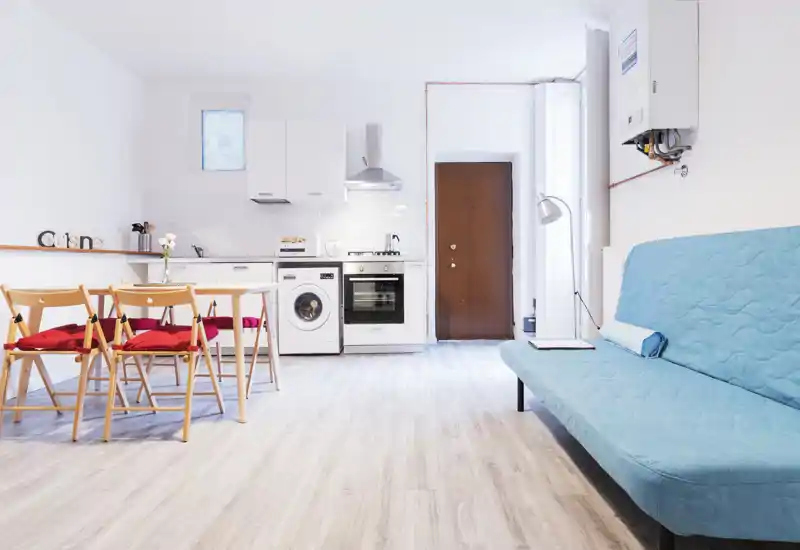 Bright, modern open-plan living area with a compact kitchen and a blue sofa. Light wood flooring and red-cushioned folding chairs give a fresh, airy feel ideal for short-term rentals.