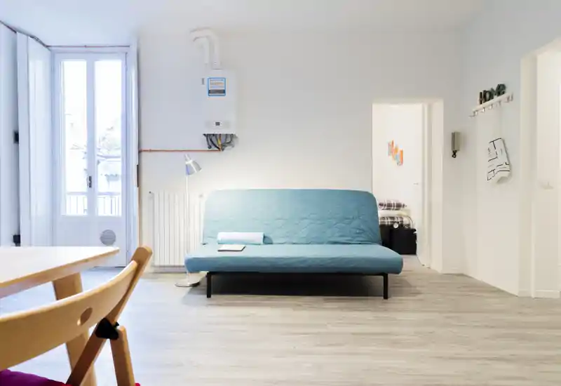 Bright, minimalist living area with a teal sofa/sofa-bed, floor lamp and large window providing abundant daylight — ideal for a compact apartment.