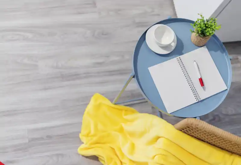 Cozy corner of a living area with a small blue side table, a notebook and pen, a cup, a potted plant and a soft yellow throw on a wicker chair — bright, tidy and inviting for relaxing or light work.