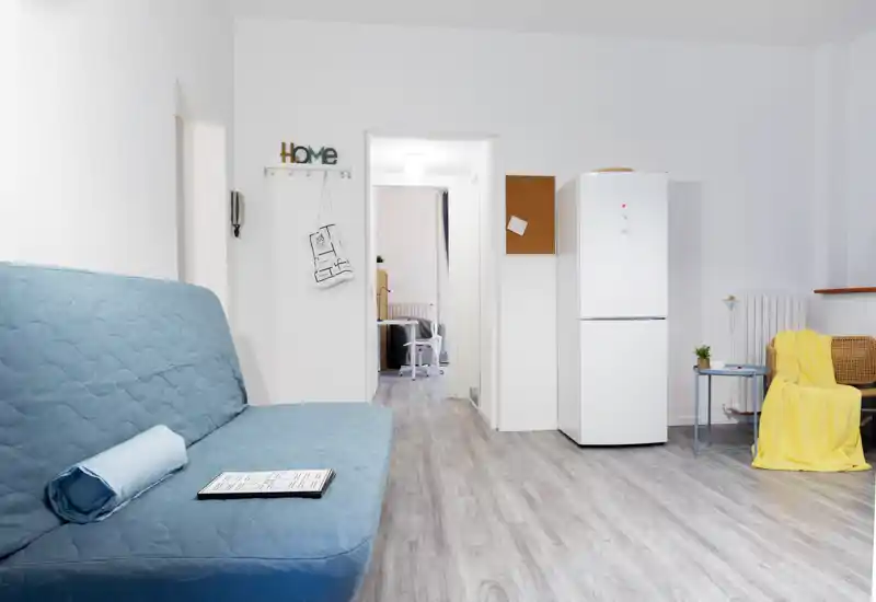 Bright, modern living area with a blue sofa, small side table and cozy yellow throw; open doorway leads to another room and a fridge sits against the wall — an inviting multifunctional living space.
