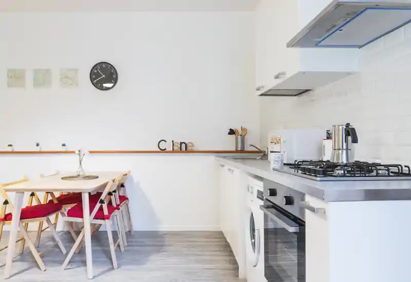 Bright modern kitchen with integrated appliances and a small dining table with red-cushioned chairs — clean white cabinetry and a stainless-steel worktop offering a contemporary, welcoming space.