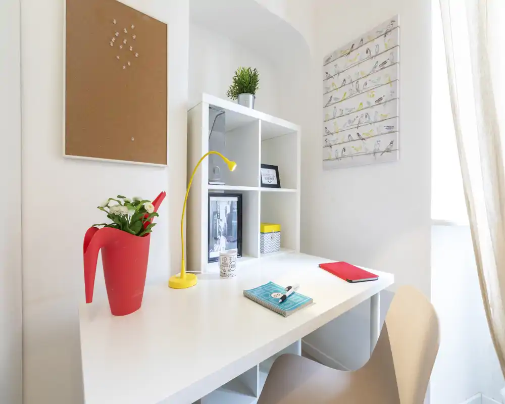 Bright, tidy workspace with a white desk, shelving unit, small decorative items and a window providing natural light — ideal for remote work or study.