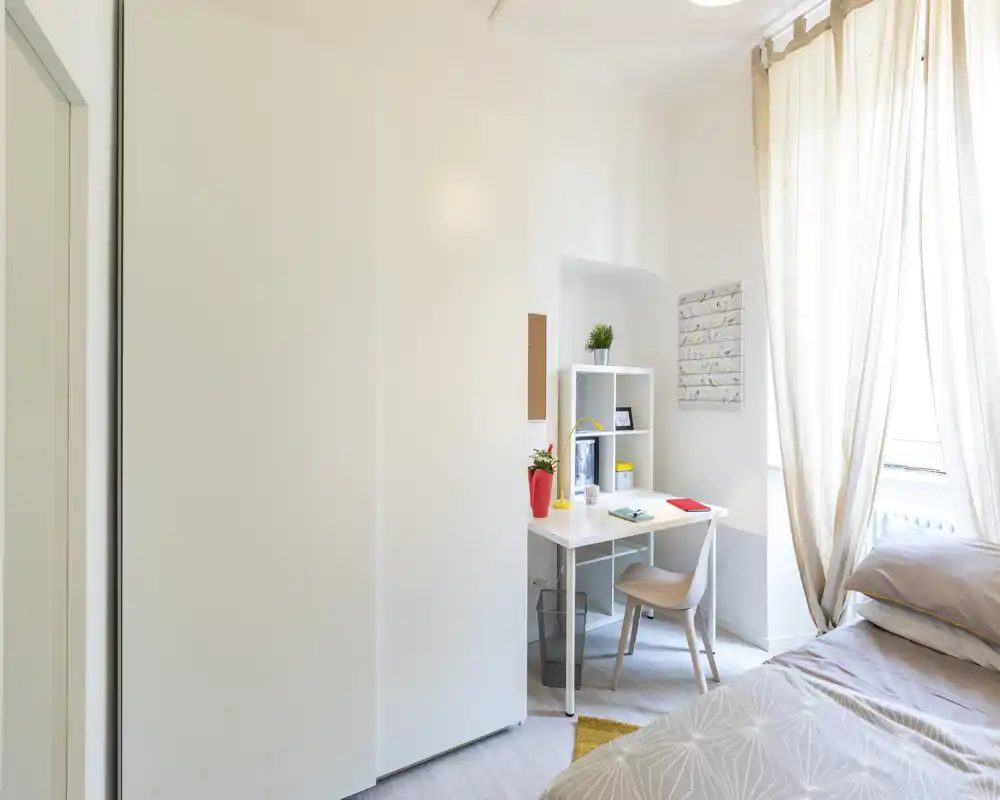 Bright, neatly styled bedroom with a comfortable bed at the right, a small desk and shelving by the window—ideal for restful stays or remote work.