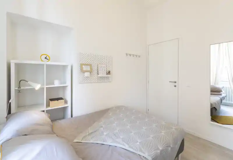 Bright, minimal bedroom with a comfortable double bed, bedside shelving and soft textiles. The room feels airy and well lit with neutral decor and a bedside lamp.