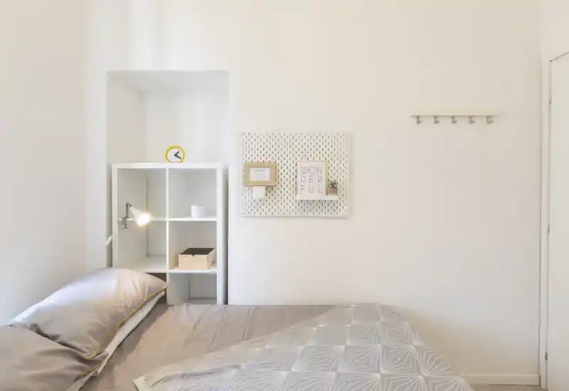 Minimal, bright bedroom with a neatly made bed, soft grey linens, a bedside shelving unit and subtle wall decor — ideal for a calm, modern rental listing.