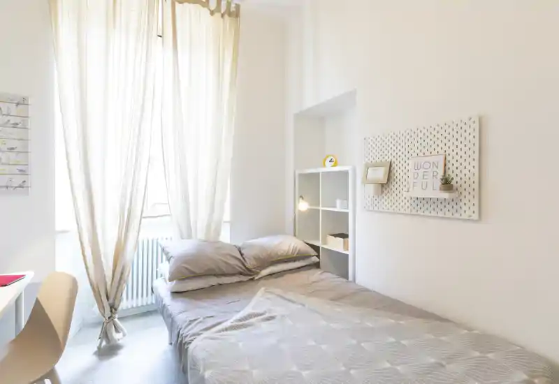 Bright and cosy single bedroom with a neatly made bed, soft pillows and neutral decor. Natural light pours through sheer curtains, highlighting minimalist shelving and tasteful wall accents.