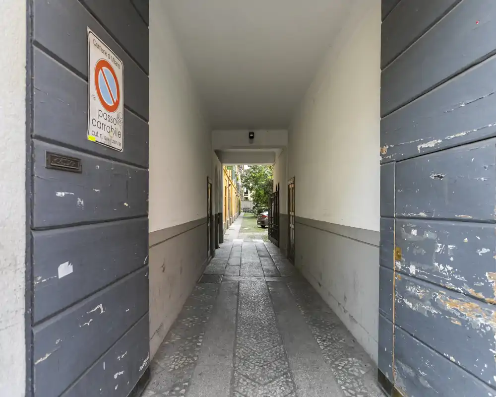 Covered entrance passage leading from the street into a courtyard, with textured stone floor and flanking doors; symmetrical, well-framed perspective.