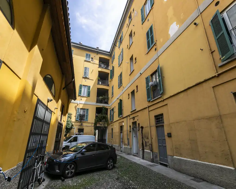 Sunny inner courtyard of a residential building with yellow façades, green shutters, parked cars and bicycles — a typical urban apartment courtyard.